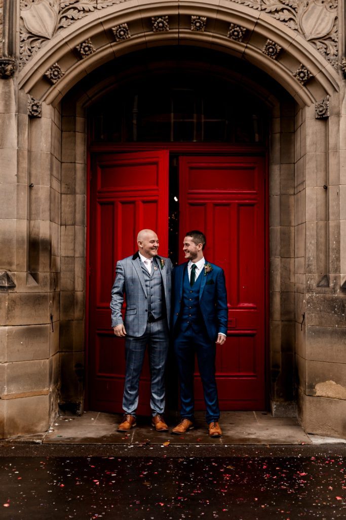 Groom & Best Man stood in front the red doon at Bakewell Town Hall. Groom is wearing a blue three-piece suit and dried flower button-hole. Photo in colour