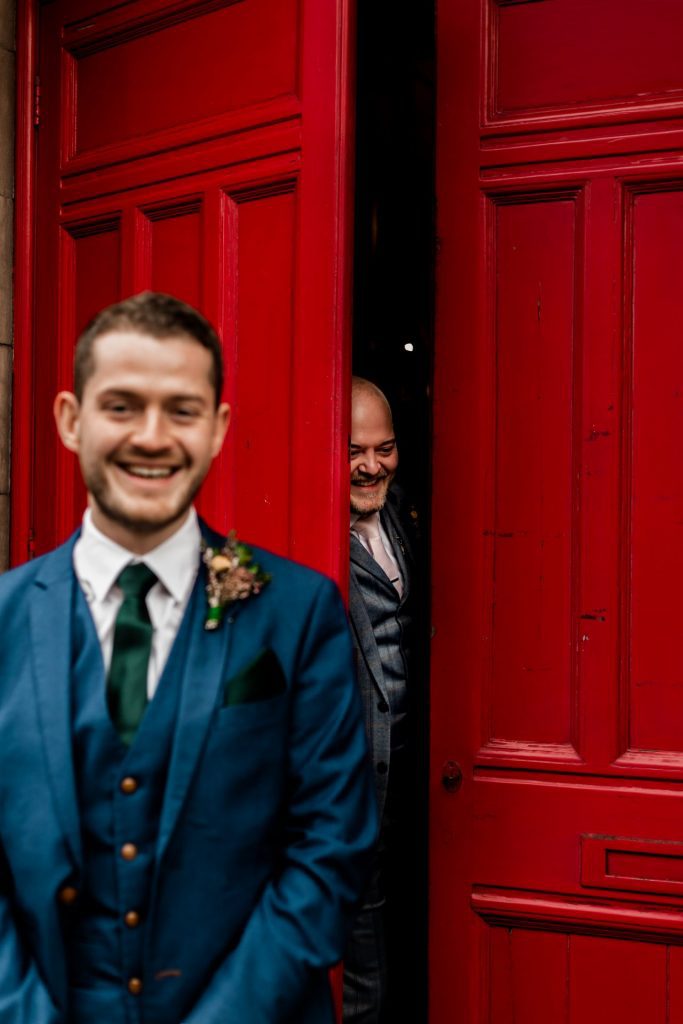 Groom stood in front the red doon at Bakewell Town Hall. Groom is wearing a blue three-piece suit and dried flower button-hole. Photo in colour