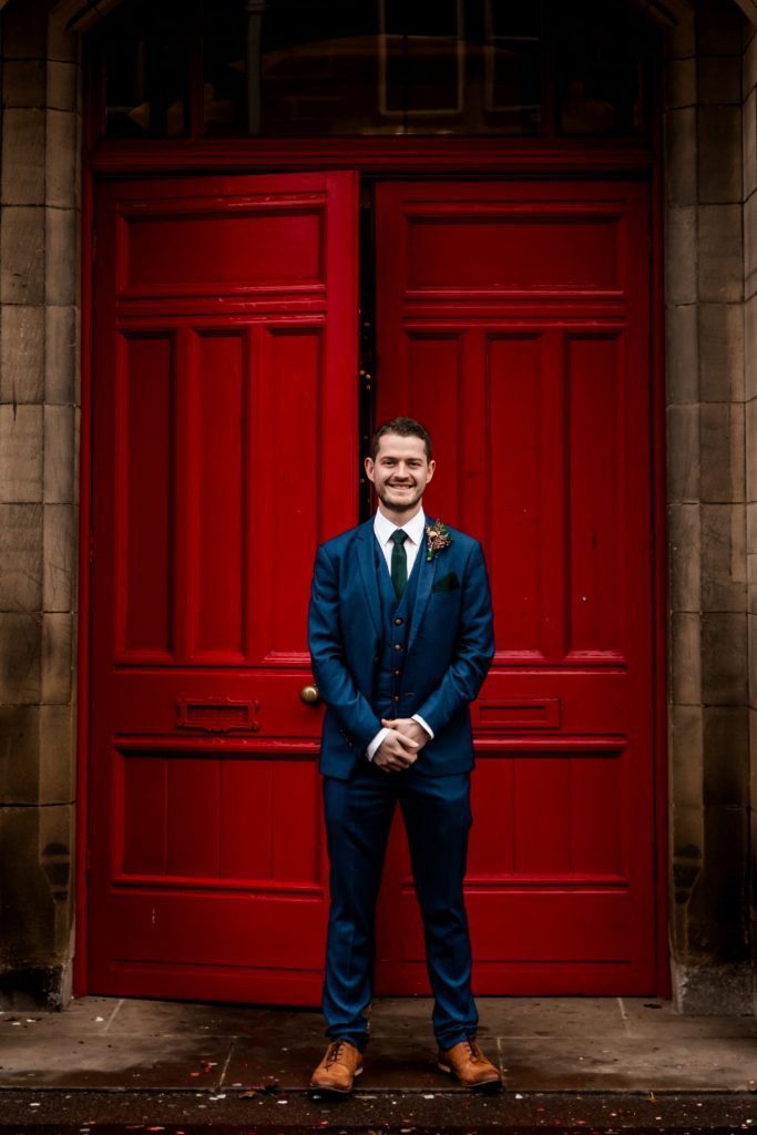 Groom stood in front the red doon at Bakewell Town Hall. Groom is wearing a blue three-piece suit and dried flower button-hole. Photo in colour