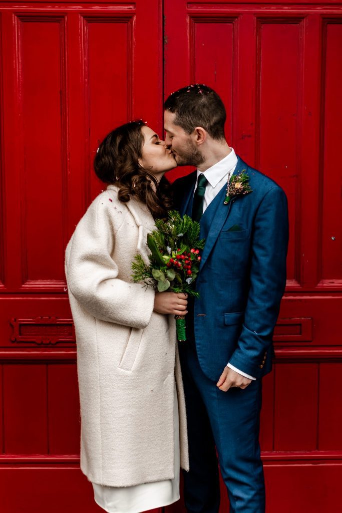 Newly married Bride and Groom stood in front the red doon at Bakewell Town Hall. Bride is wearing a white three-quarter length wedding dress with white wool coat. Groom is wearing a blue three-piece suit and dried flower button-hole. Photo in colour