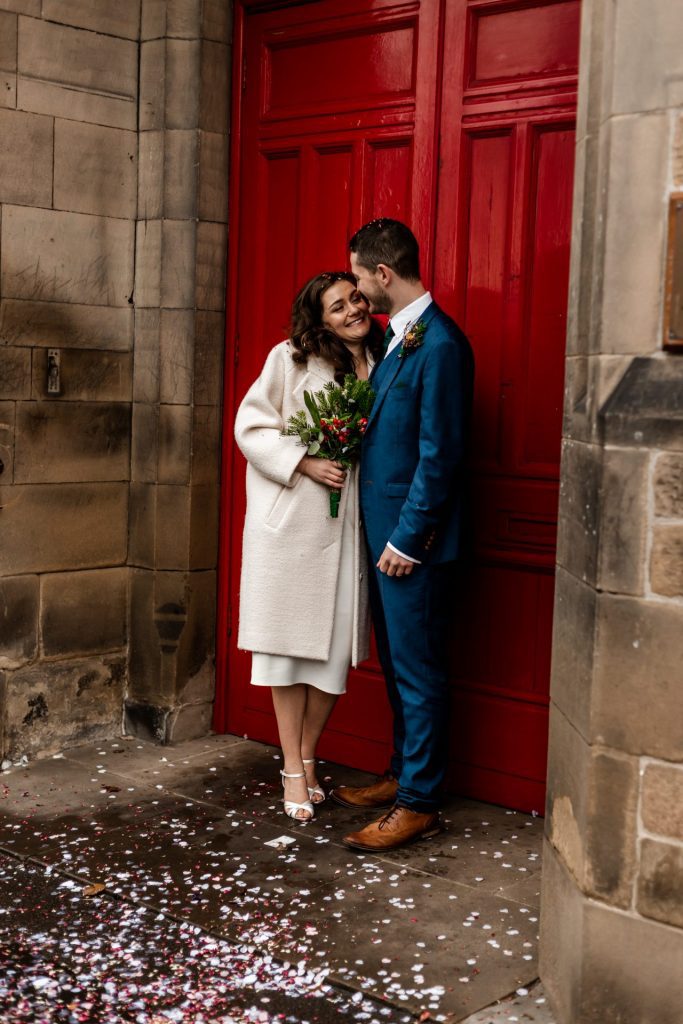 Newly married Bride and Groom stood in front the red doon at Bakewell Town Hall. Bride is wearing a white three-quarter length wedding dress with white wool coat. Groom is wearing a blue three-piece suit and dried flower button-hole. Photo in colour