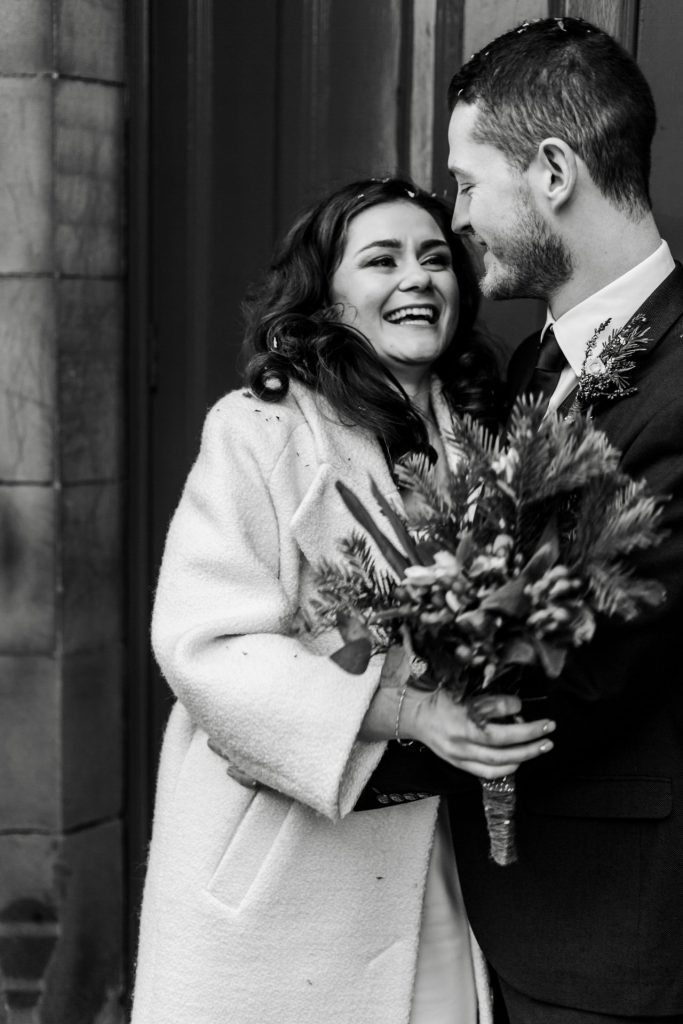 Newly married Bride and Groom stood in front the red doon at Bakewell Town Hall. Bride is wearing a white three-quarter length wedding dress with white wool coat. Groom is wearing a blue three-piece suit and dried flower button-hole. Photo in black & white