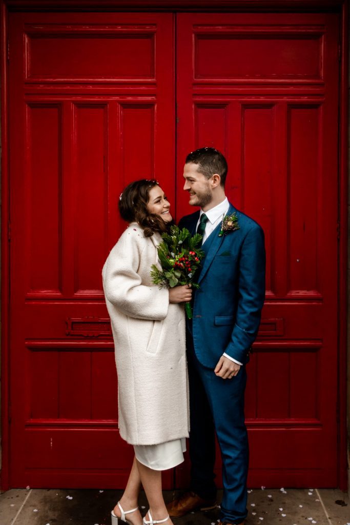 Newly married Bride and Groom stood in front the red doon at Bakewell Town Hall. Bride is wearing a white three-quarter length wedding dress with white wool coat. Groom is wearing a blue three-piece suit and dried flower button-hole. Photo in colour
