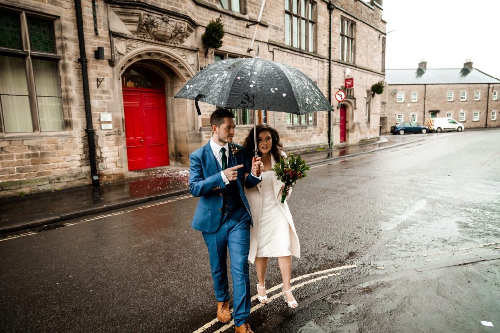Newly married Bride and Groom walking through Bakwell under a black umbrella. Bride is wearing a white three-quarter length wedding dress with white wool coat. Groom is wearing a blue three-piece suit and dried flower button-hole. Photo in colour