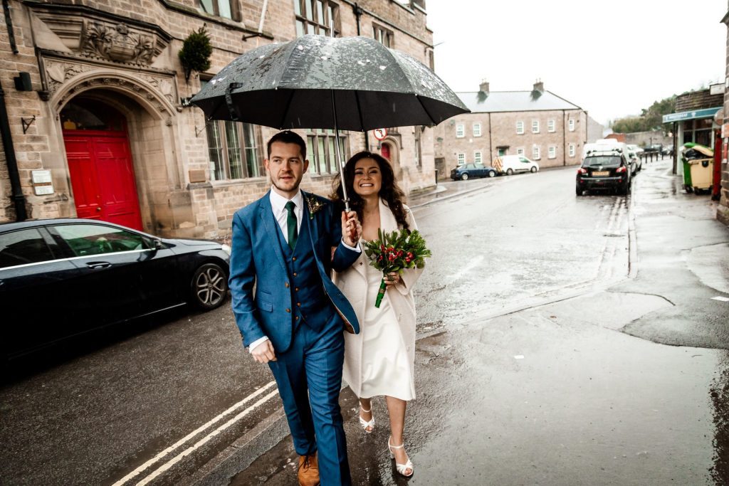 Newly married Bride and Groom walking through Bakwell under a black umbrella. Bride is wearing a white three-quarter length wedding dress with white wool coat. Groom is wearing a blue three-piece suit and dried flower button-hole. Photo in colour