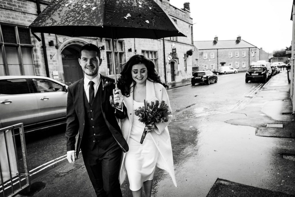 Newly married Bride and Groom walking through Bakwell under a black umbrella. Bride is wearing a white three-quarter length wedding dress with white wool coat. Groom is wearing a blue three-piece suit and dried flower button-hole. Photo in black & white