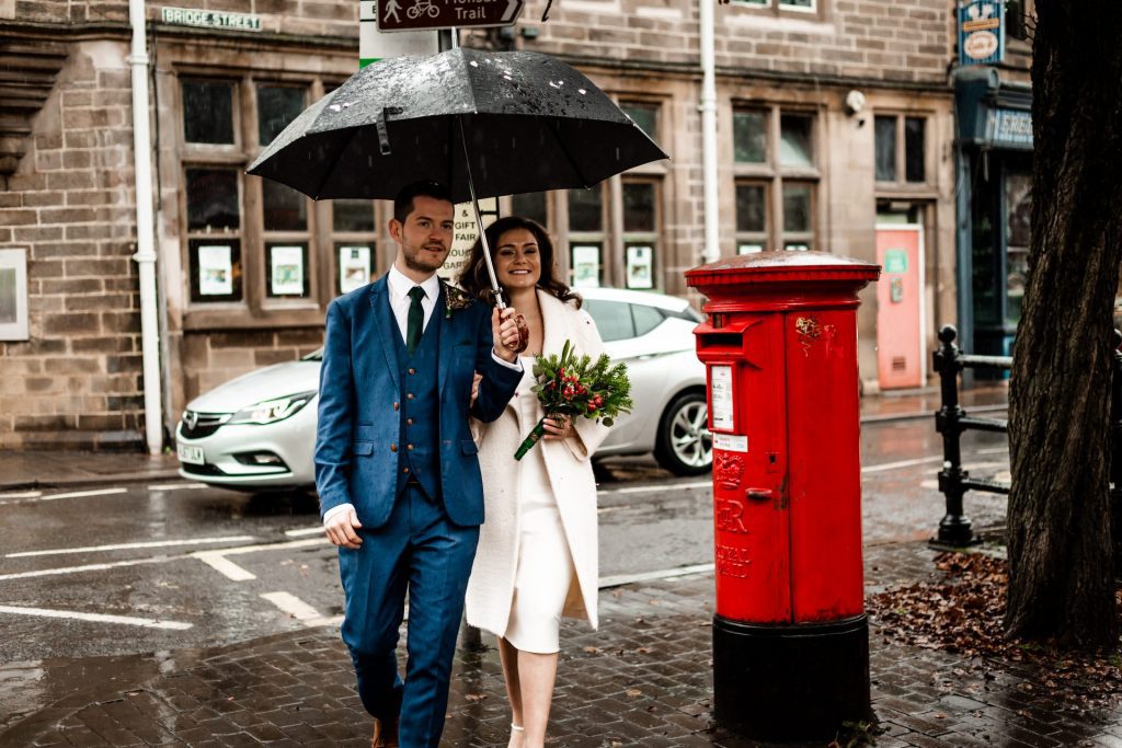Newly married Bride and Groom walking through Bakwell under a black umbrella. Bride is wearing a white three-quarter length wedding dress with white wool coat. Groom is wearing a blue three-piece suit and dried flower button-hole. Photo in colour