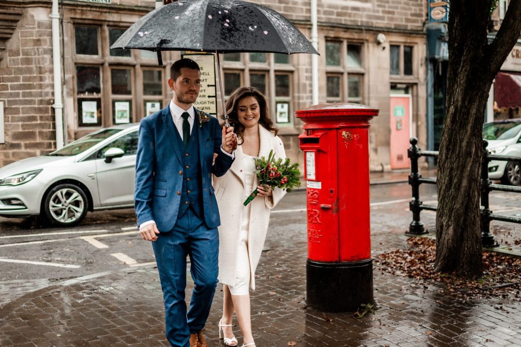 Newly married Bride and Groom walking through Bakwell under a black umbrella. Bride is wearing a white three-quarter length wedding dress with white wool coat. Groom is wearing a blue three-piece suit and dried flower button-hole. Photo in colour