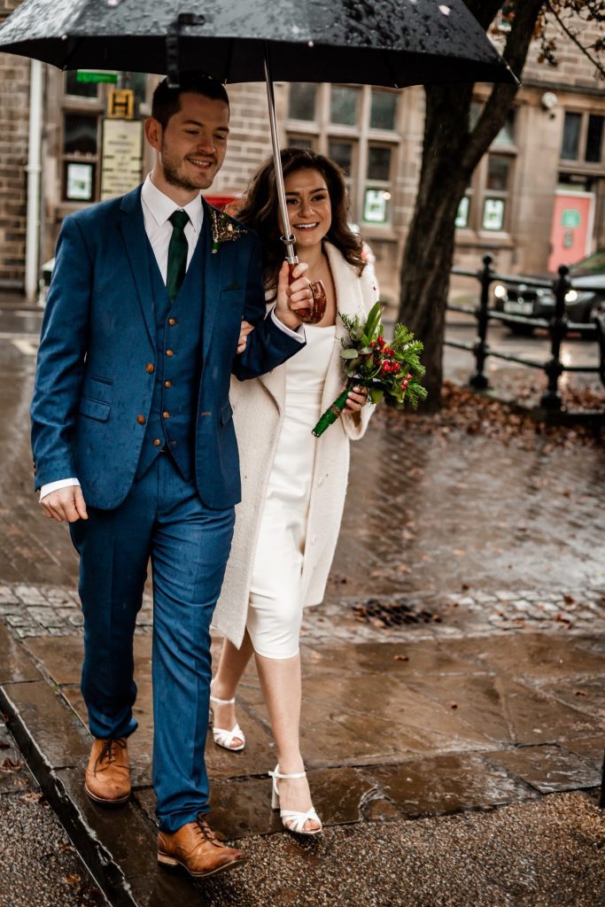 Newly married Bride and Groom walking through Bakwell under a black umbrella. Bride is wearing a white three-quarter length wedding dress with white wool coat. Groom is wearing a blue three-piece suit and dried flower button-hole. Photo in colour