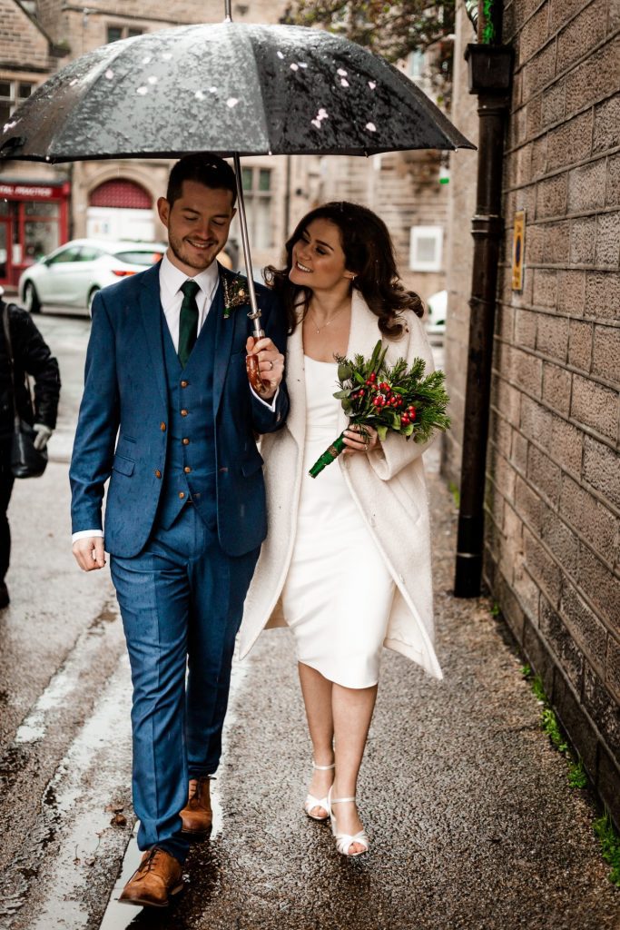 Newly married Bride and Groom walking through Bakwell under a black umbrella. Bride is wearing a white three-quarter length wedding dress with white wool coat. Groom is wearing a blue three-piece suit and dried flower button-hole. Photo in colour