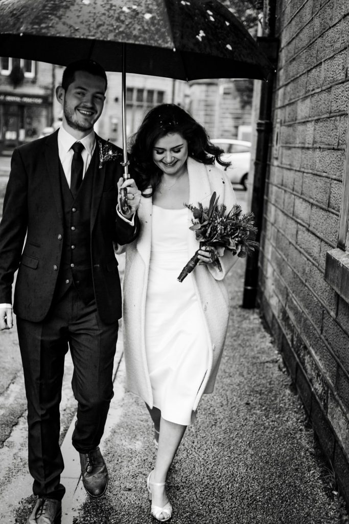 Newly married Bride and Groom walking through Bakwell under a black umbrella. Bride is wearing a white three-quarter length wedding dress with white wool coat. Groom is wearing a blue three-piece suit and dried flower button-hole. Photo in black & white
