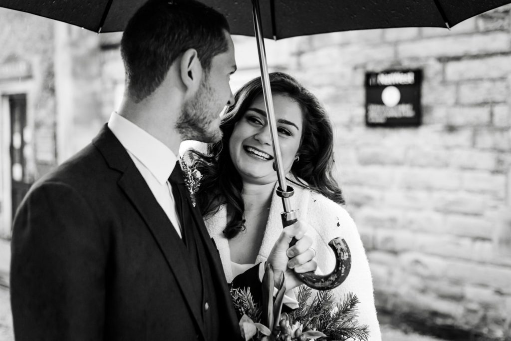 Newly married Bride and Groom walking through Bakwell under a black umbrella. Bride is wearing a white three-quarter length wedding dress with white wool coat. Groom is wearing a blue three-piece suit and dried flower button-hole. Photo in black & white