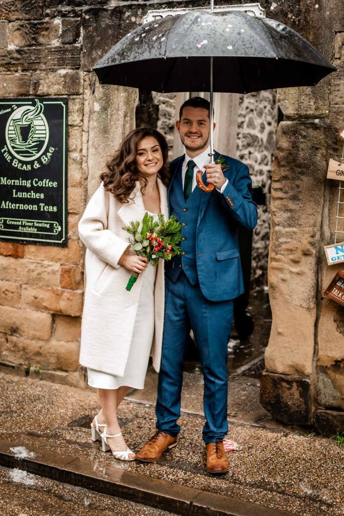 Newly married Bride and Groom walking through Bakwell under a black umbrella. Bride is wearing a white three-quarter length wedding dress with white wool coat. Groom is wearing a blue three-piece suit and dried flower button-hole. Photo in colour