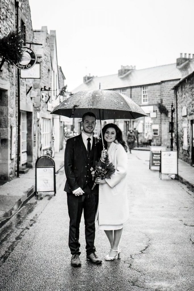 Newly married Bride and Groom walking through Bakwell under a black umbrella. Bride is wearing a white three-quarter length wedding dress with white wool coat. Groom is wearing a blue three-piece suit and dried flower button-hole. Photo in black & white