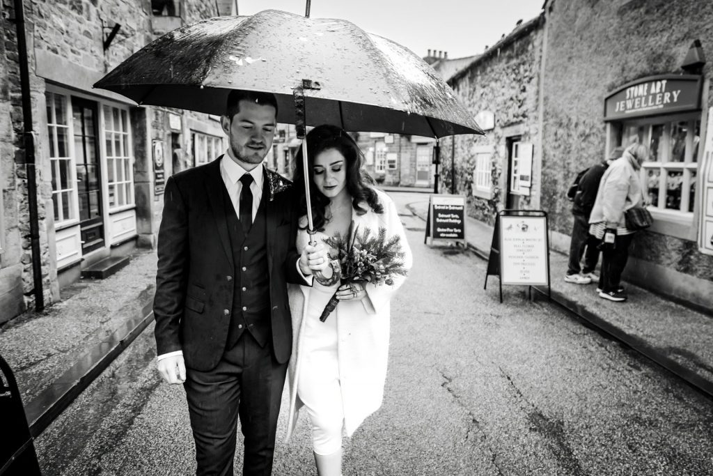 Newly married Bride and Groom walking through Bakwell under a black umbrella. Bride is wearing a white three-quarter length wedding dress with white wool coat. Groom is wearing a blue three-piece suit and dried flower button-hole. Photo in black & white