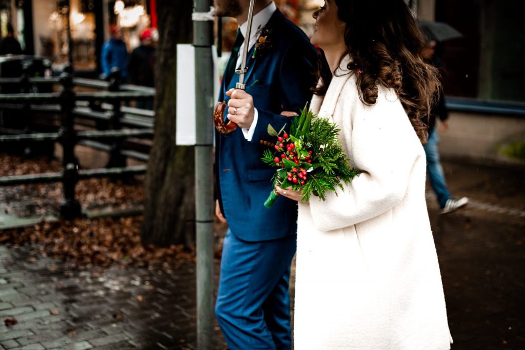Newly married Bride and Groom walking through Bakwell under a black umbrella. Bride is wearing a white three-quarter length wedding dress with white wool coat. Groom is wearing a blue three-piece suit and dried flower button-hole. Photo in colour