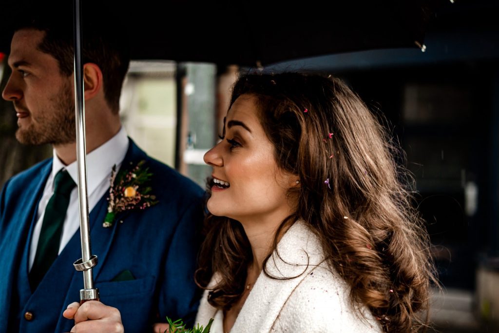 Newly married Bride and Groom walking through Bakwell under a black umbrella. Bride is wearing a white three-quarter length wedding dress with white wool coat. Groom is wearing a blue three-piece suit and dried flower button-hole. Photo in colour
