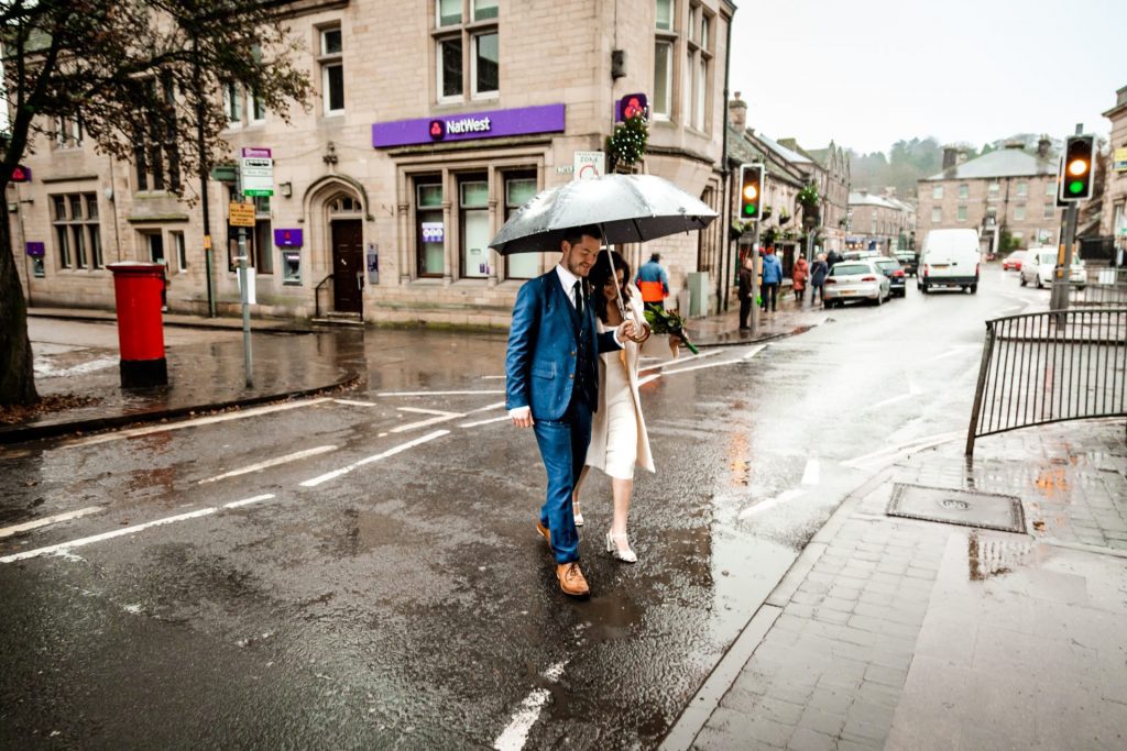 Newly married Bride and Groom walking through Bakwell under a black umbrella. Bride is wearing a white three-quarter length wedding dress with white wool coat. Groom is wearing a blue three-piece suit and dried flower button-hole. Photo in colour