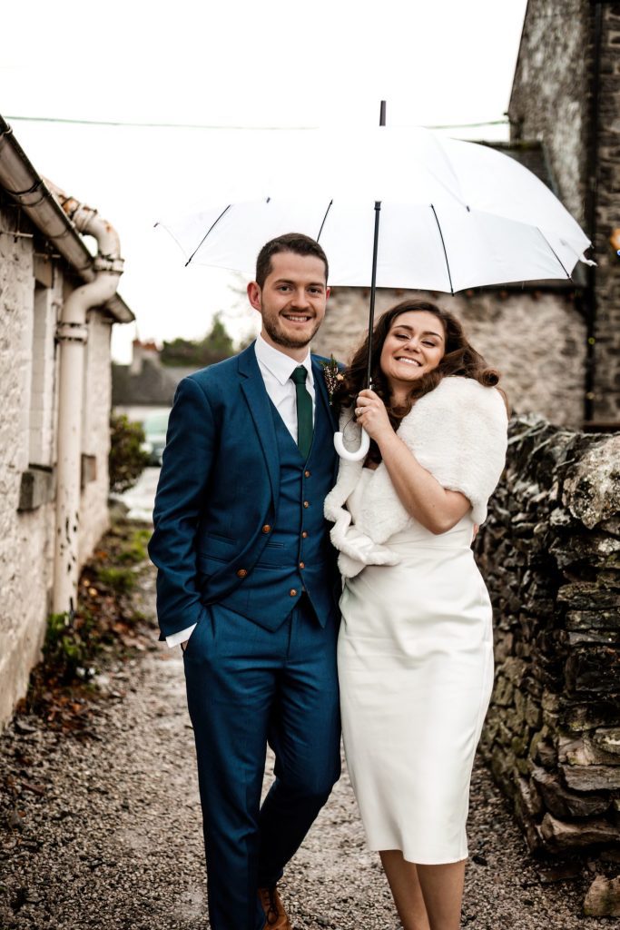 Newly married Bride and Groom cuddling together under a white umbrella at the Monsall Head Pub. Bride is wearing a white three-quarter length dress and faux-fur shawl. Groom is wearing a blue three-piece suit and dried flower button-hole. Cropped in. Photo in colour