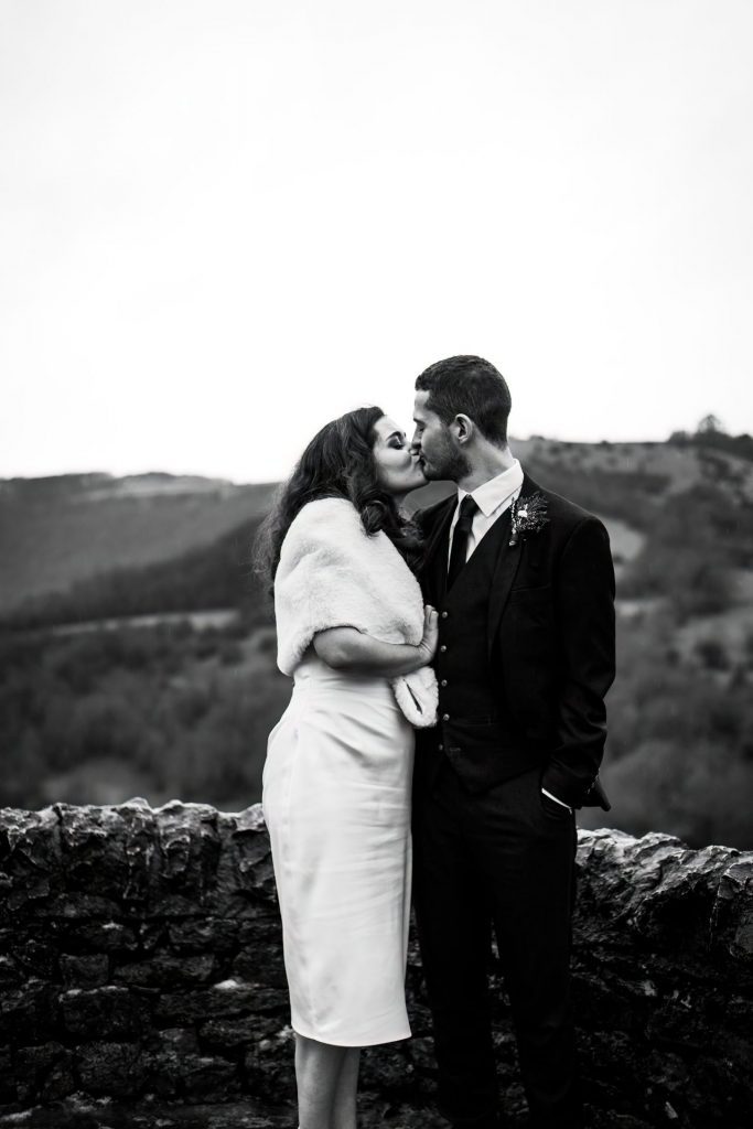 Newly married Bride and Groom cuddling together with the view of Monsall Head behind them. Bride is wearing a white three-quarter length dress and faux-fur shawl. Groom is wearing a blue three-piece suit and dried flower button-hole. Cropped in. Photo in black & white