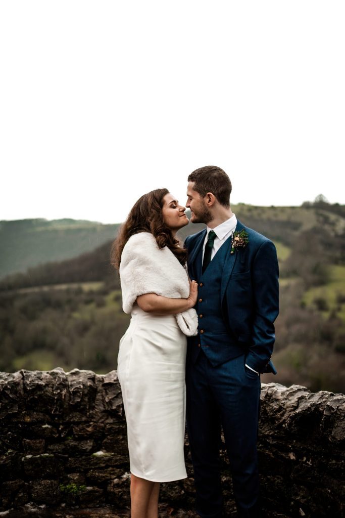 Newly married Bride and Groom cuddling together with the view of Monsall Head behind them. Bride is wearing a white three-quarter length dress and faux-fur shawl. Groom is wearing a blue three-piece suit and dried flower button-hole. Cropped in. Photo in colour
