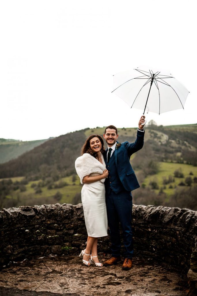 Newly married Bride and Groom cuddling together under a white umbrella with the view of Monsall Head behind them. Bride is wearing a white three-quarter length dress and faux-fur shawl. Groom is wearing a blue three-piece suit and dried flower button-hole. Cropped in. Photo in colour