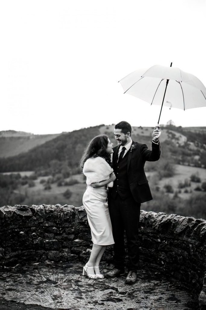 Newly married Bride and Groom cuddling together under a white umbrella with the view of Monsall Head behind them. Bride is wearing a white three-quarter length dress and faux-fur shawl. Groom is wearing a blue three-piece suit and dried flower button-hole. Cropped in. Photo in black & white