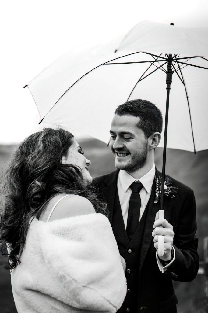 Close up of newly married Bride and Groom cuddling together under a white umbrella with the view of Monsall Head behind them. Bride is wearing a white three-quarter length dress and faux-fur shawl. Groom is wearing a blue three-piece suit and dried flower button-hole. Cropped in. Photo in black & white