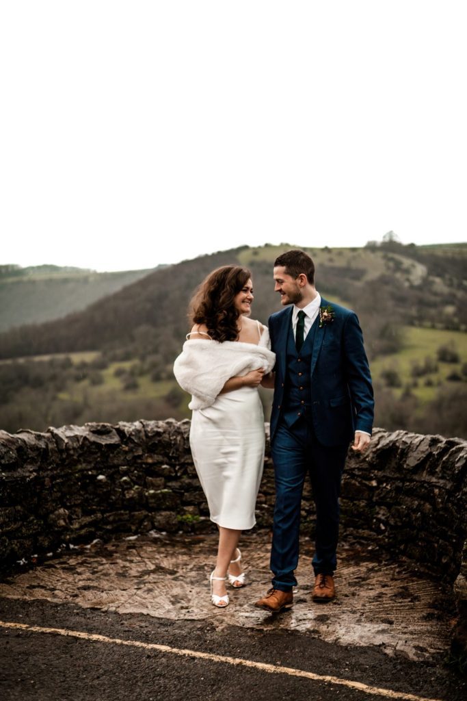 Newly married Bride and Groom cuddle together with the view of Monsall Head behind them. Bride is wearing a white three-quarter length dress and faux-fur shawl. Groom is wearing a blue three-piece suit and dried flower button-hole. Cropped in. Photo in colour