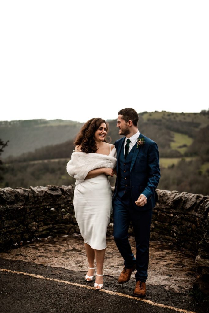 Newly married Bride and Groom cuddle together with the view of Monsall Head behind them. Bride is wearing a white three-quarter length dress and faux-fur shawl. Groom is wearing a blue three-piece suit and dried flower button-hole. Cropped in. Photo in colour