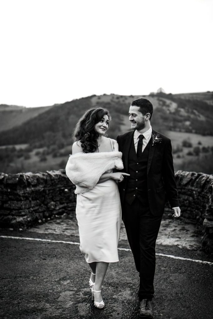 Newly married Bride and Groom cuddle together with the view of Monsall Head behind them. Bride is wearing a white three-quarter length dress and faux-fur shawl. Groom is wearing a blue three-piece suit and dried flower button-hole. Cropped in. Photo in black & white