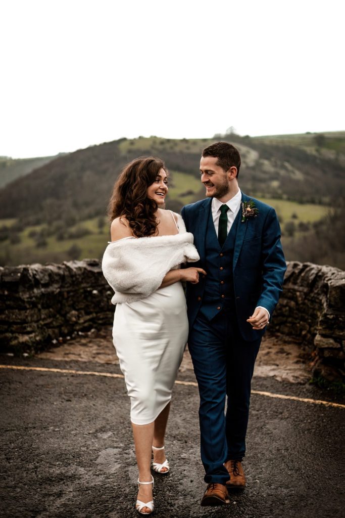 Newly married Bride and Groom cuddle together with the view of Monsall Head behind them. Bride is wearing a white three-quarter length dress and faux-fur shawl. Groom is wearing a blue three-piece suit and dried flower button-hole. Cropped in. Photo in colour