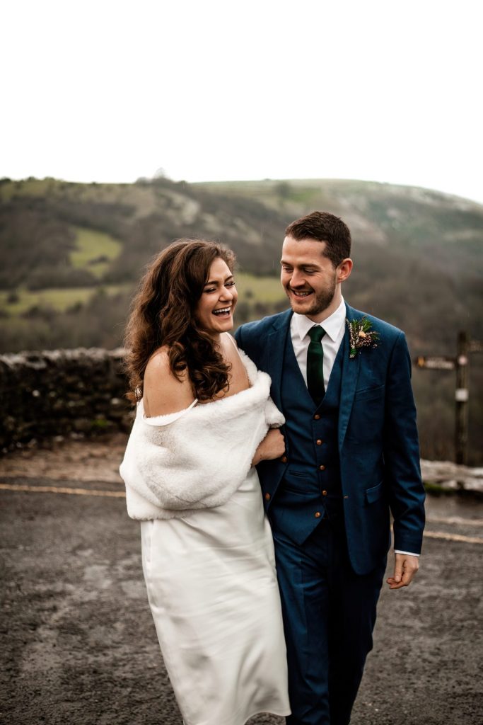 Newly married Bride and Groom cuddle together with the view of Monsall Head behind them. Bride is wearing a white three-quarter length dress and faux-fur shawl. Groom is wearing a blue three-piece suit and dried flower button-hole. Cropped in. Photo in colour