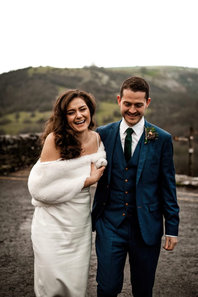 Newly married Bride and Groom cuddle together with the view of Monsall Head behind them. Bride is wearing a white three-quarter length dress and faux-fur shawl. Groom is wearing a blue three-piece suit and dried flower button-hole. Cropped in. Photo in colour