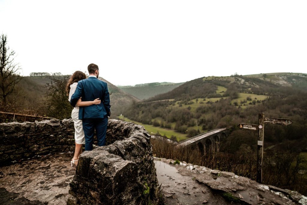 Newly married Bride and Groom cuddle together with their back turned looking towards the view of Monsall Head behind them. Bride is wearing a white three-quarter length dress and faux-fur shawl. Groom is wearing a blue three-piece suit and dried flower button-hole. Cropped in. Photo in colour