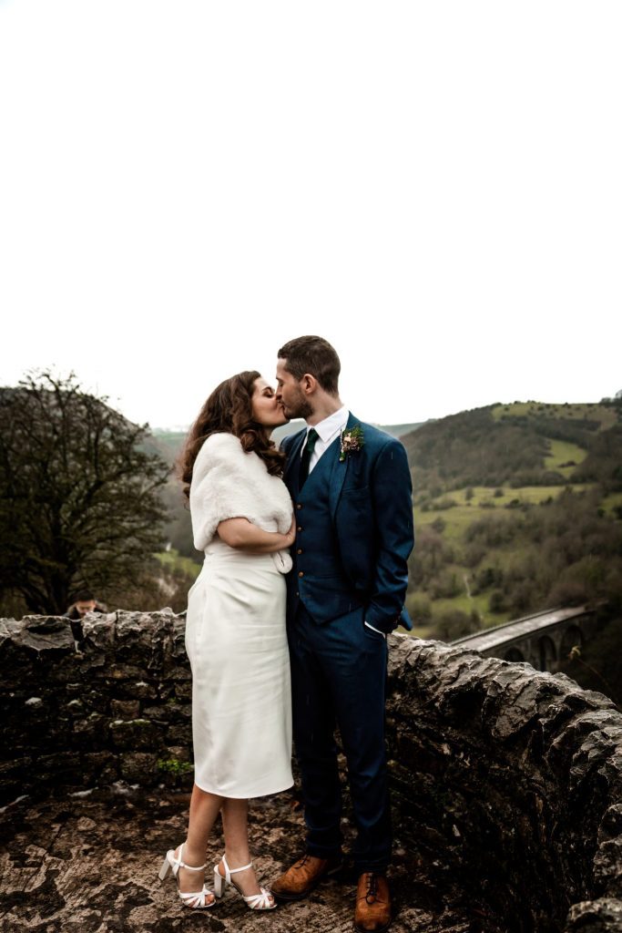 Newly married Bride and Groom kissing with the view of Monsall Head behind them. Bride is wearing a white three-quarter length dress and faux-fur shawl. Groom is wearing a blue three-piece suit and dried flower button-hole. Cropped in. Photo in colour