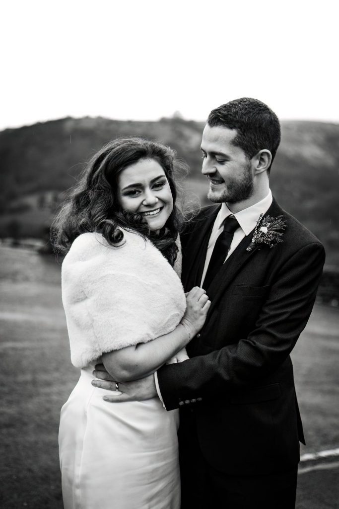 Newly married Bride and Groom cuddle together with the view of Monsall Head behind them. Bride is wearing a white three-quarter length dress and faux-fur shawl. Groom is wearing a blue three-piece suit and dried flower button-hole. Cropped in. Photo in black & white