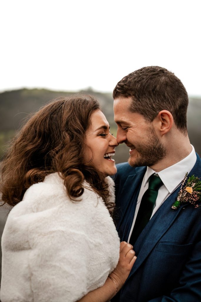Newly married Bride and Groom cuddle together with the view of Monsall Head behind them. Close in shot of the Bride smiling. Bride is wearing a white three-quarter length dress and faux-fur shawl. Groom is wearing a blue three-piece suit and dried flower button-hole. Photo in colour