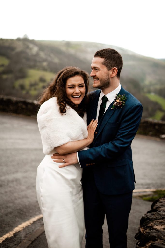 Newly married Bride and Groom cuddle together with the view of Monsall Head behind them. Bride is wearing a white three-quarter length dress and faux-fur shawl. Groom is wearing a blue three-piece suit and dried flower button-hole. Photo in colour