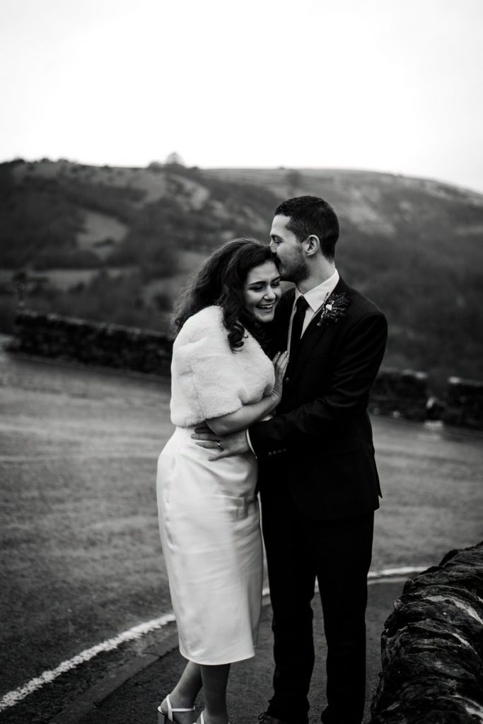 Newly married Bride and Groom cuddle together with the view of Monsall Head behind them. Bride is wearing a white three-quarter length dress and faux-fur shawl. Groom is wearing a blue three-piece suit and dried flower button-hole. Photo in black & white