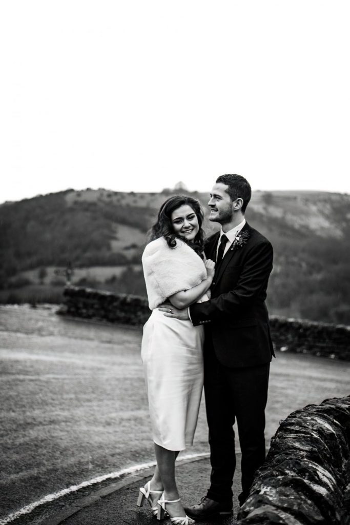 Newly married Bride and Groom cuddle together with the view of Monsall Head behind them. Bride is wearing a white three-quarter length dress and faux-fur shawl. Groom is wearing a blue three-piece suit and dried flower button-hole. Photo in black & white