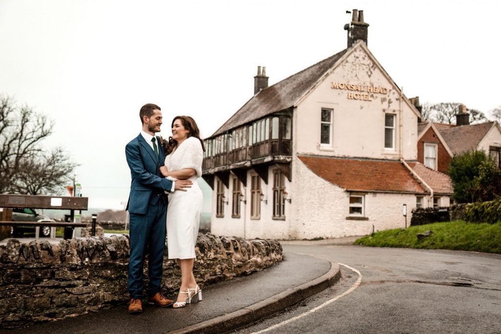 Newly married Bride and Groom in front the Monsall Head Pub. Bride is wearing a white three-quarter length dress and faux-fur shawl. Groom is wearing a blue three-piece suit and dried flower button-hole. Photo in colour
