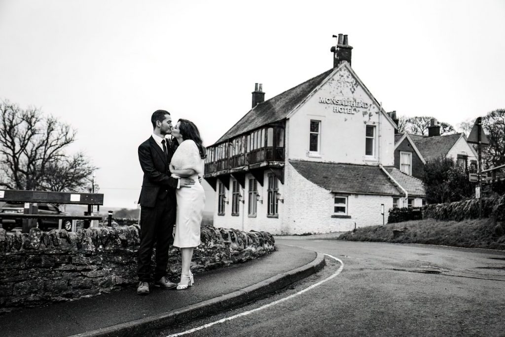 Newly married Bride and Groom kissing in front the Monsall Head Pub. Bride is wearing a white three-quarter length dress and faux-fur shawl. Groom is wearing a blue three-piece suit and dried flower button-hole. Photo in black & white