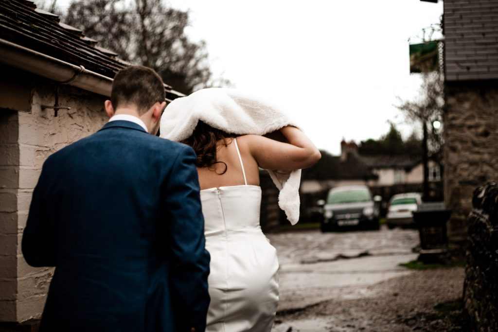 Candid photo of ewly married Bride & Groom sheltering from the rain using the Brides Shawl. Bride is wearing a white three-quarter length dress and faux-fur shawl. Photo in colour