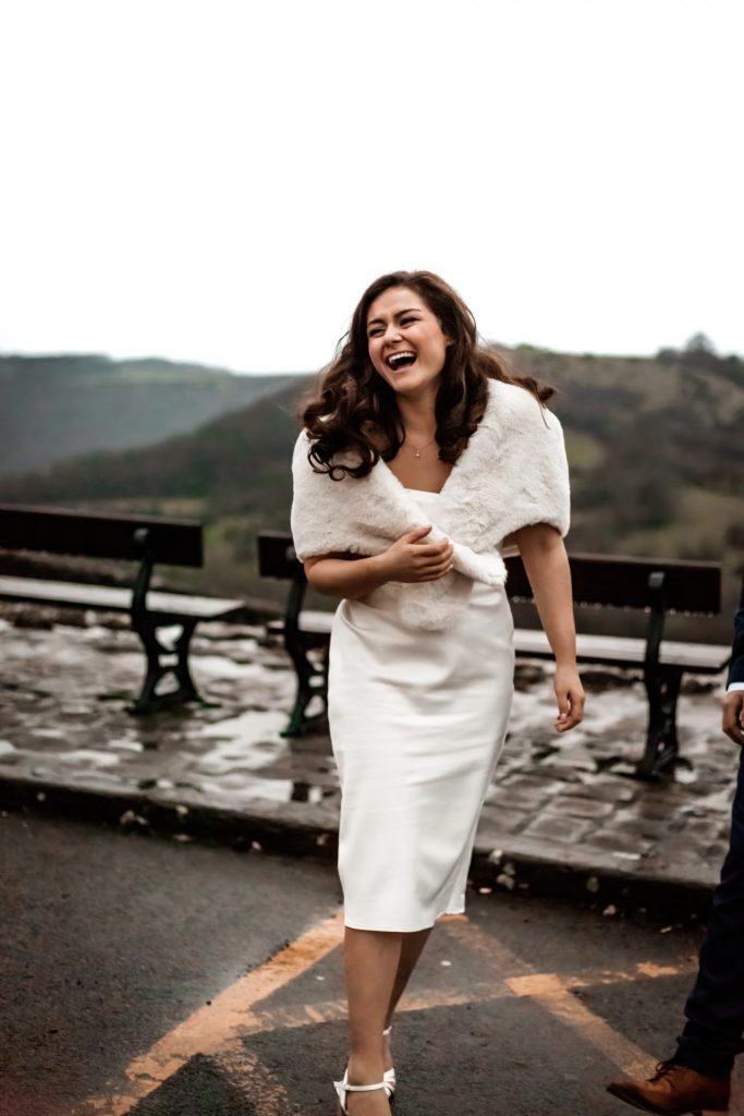 Newly married Bride caught laughing at Monsall Head Pub. Bride is wearing a white three-quarter length dress and faux-fur shawl. Photo in colour