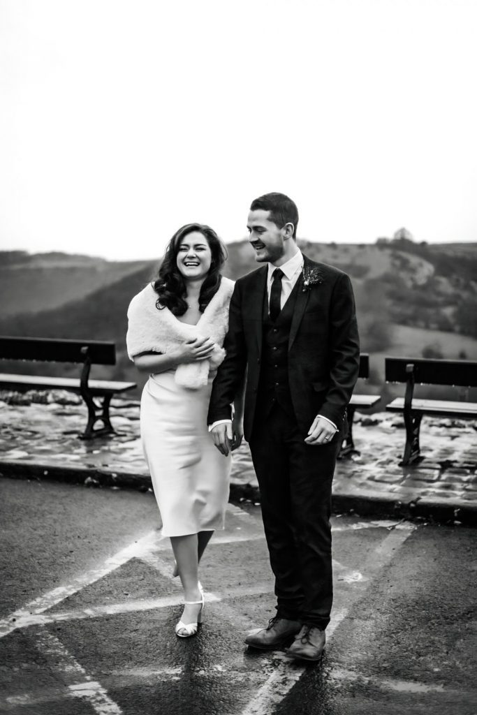 Newly married Bride and Groom take stroll at Monsall Head Pub. Bride is wearing a white three-quarter length dress and faux-fur shawl. Groom is wearing a blue three-piece suit and dried flower button-hole. Photo in black and white