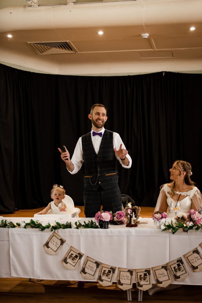 Groom stood delivering his speech in a waistcoat and purple bowtie as his new wife watches smiling