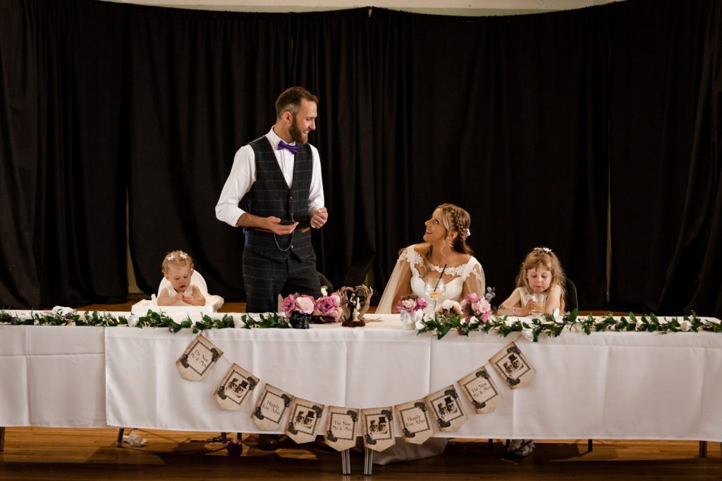 Groom stood delivering his speech in a waistcoat and purple bowtie as his new wife watches smiling