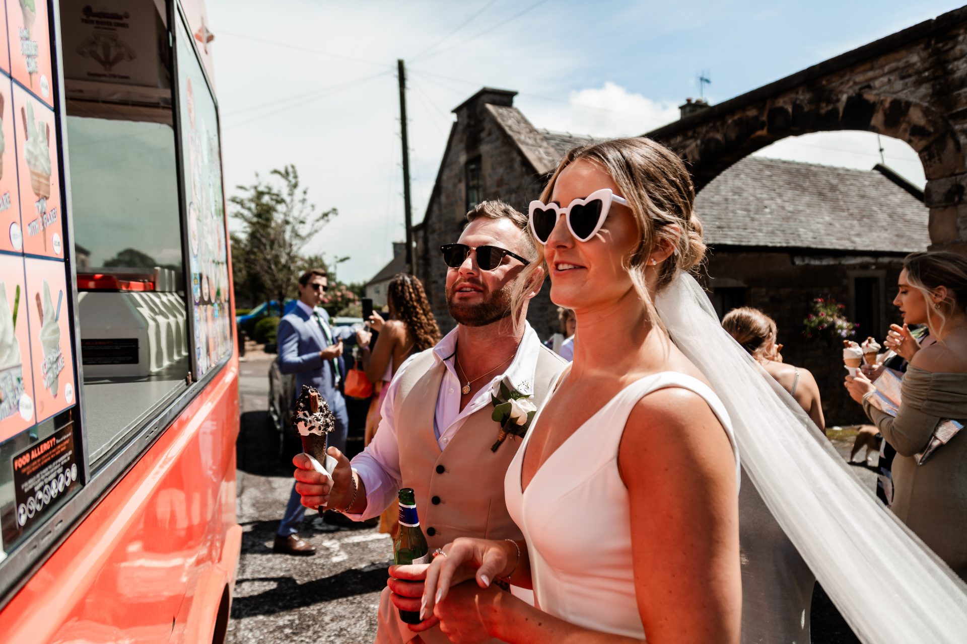Close up shot of the newly married bride & groom waiting to get an ice cream from a ice cream van at their wedding venue. The bride is wearing white heart sunglasses