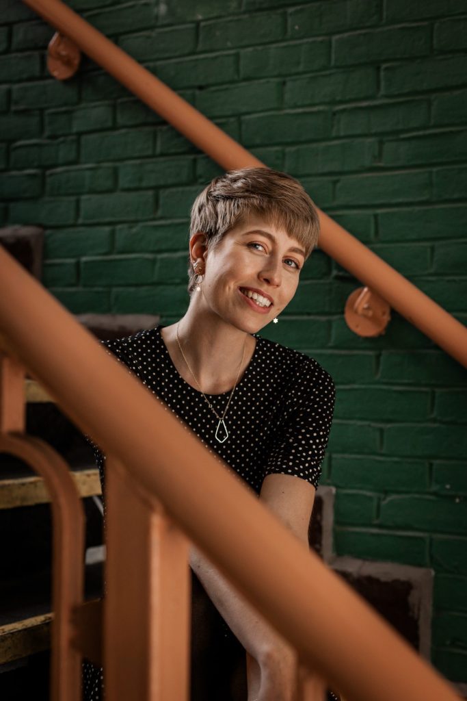 Isabelle Biggs wedding photographer pictured sitting on a staircase with a green painted brick wall in the background